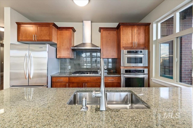 kitchen featuring sink, stainless steel appliances, wall chimney range hood, light stone counters, and backsplash