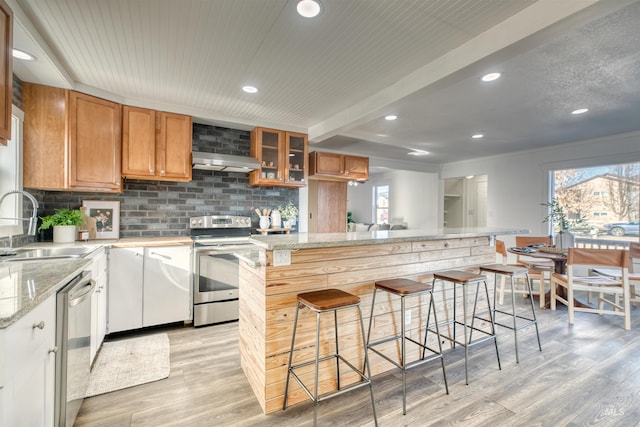 kitchen with exhaust hood, sink, appliances with stainless steel finishes, light stone counters, and a breakfast bar area