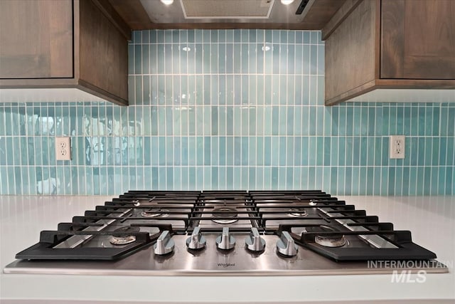 kitchen view of stainless steel gas cooktop, backsplash, dark brown cabinets, and range hood