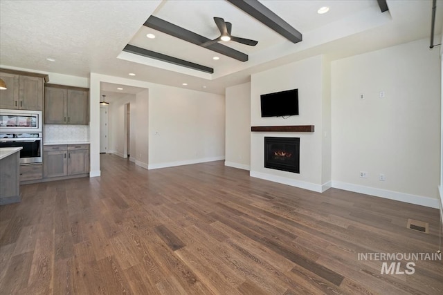 unfurnished living room with ceiling fan, dark wood-style flooring, a tray ceiling, a lit fireplace, and baseboards