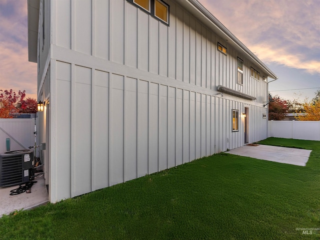 property exterior at dusk featuring fence, a lawn, a patio area, central AC unit, and board and batten siding