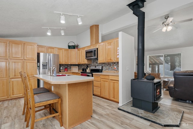 kitchen with a wood stove, light brown cabinets, tasteful backsplash, a center island with sink, and appliances with stainless steel finishes