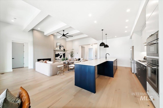 kitchen featuring beam ceiling, decorative light fixtures, a center island with sink, appliances with stainless steel finishes, and light wood-type flooring