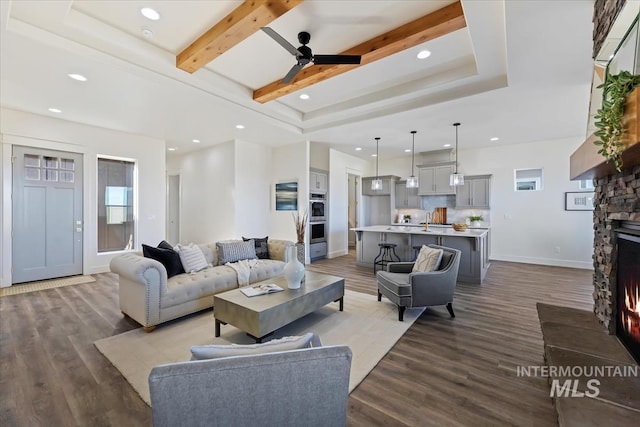 living room featuring wood-type flooring, beamed ceiling, a stone fireplace, sink, and ceiling fan