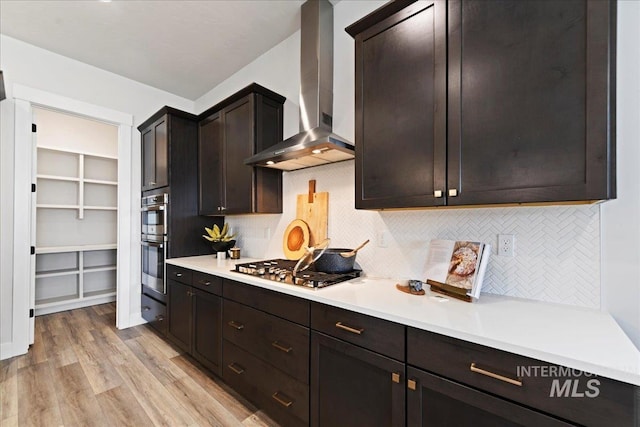 kitchen with decorative backsplash, wall chimney exhaust hood, dark brown cabinetry, stainless steel appliances, and light hardwood / wood-style flooring