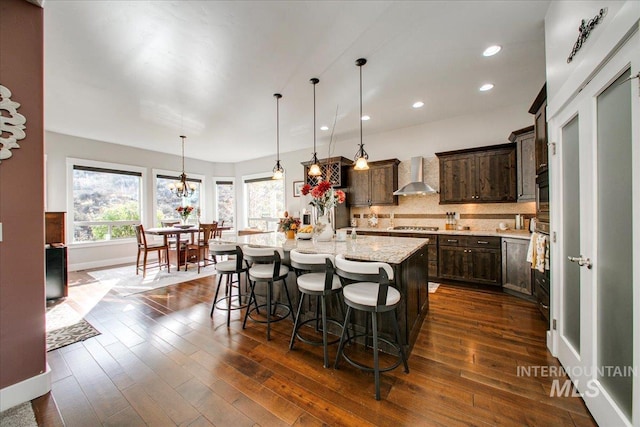 kitchen with dark wood-type flooring, wall chimney range hood, a center island with sink, and hanging light fixtures