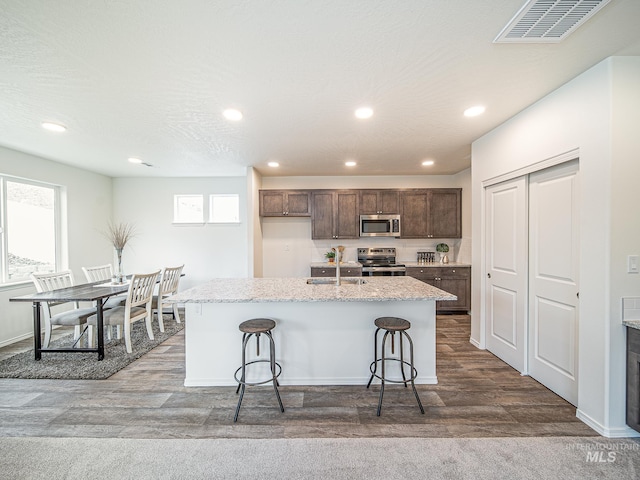 kitchen with hardwood / wood-style floors, a kitchen bar, a kitchen island with sink, light stone counters, and stainless steel appliances