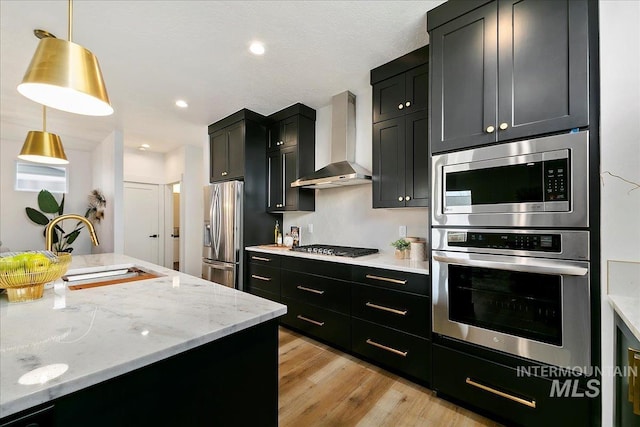 kitchen featuring wall chimney exhaust hood, hanging light fixtures, light stone counters, appliances with stainless steel finishes, and light wood-type flooring