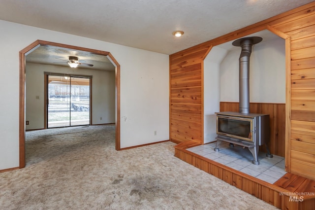 unfurnished living room featuring wooden walls, carpet floors, a wood stove, arched walkways, and a ceiling fan