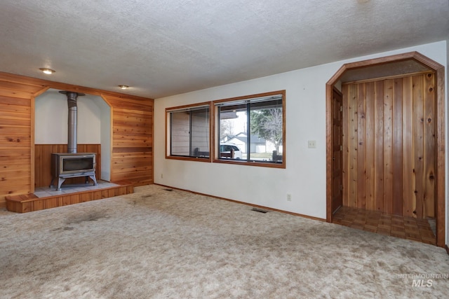 unfurnished living room featuring carpet floors, wooden walls, a wood stove, and a textured ceiling