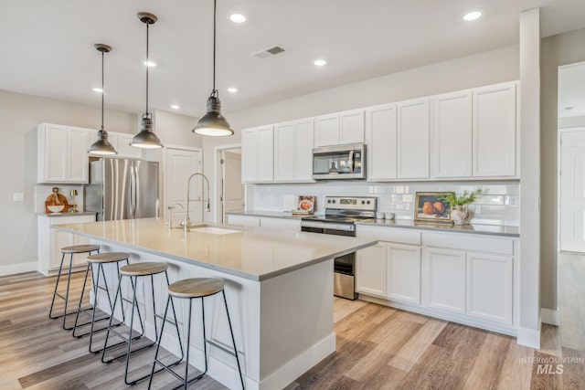 kitchen with stainless steel appliances, decorative light fixtures, white cabinetry, light hardwood / wood-style floors, and a center island with sink