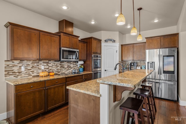 kitchen with dark wood-type flooring, a center island with sink, hanging light fixtures, appliances with stainless steel finishes, and light stone counters