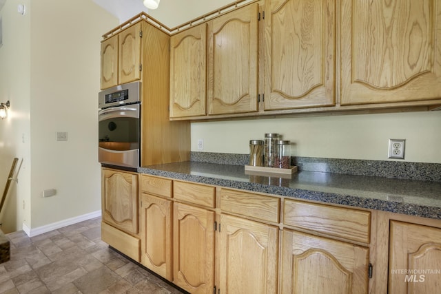 kitchen featuring oven, dark countertops, baseboards, stone finish flooring, and light brown cabinets