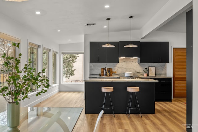 kitchen with a kitchen breakfast bar, light wood-type flooring, dark cabinetry, decorative backsplash, and light countertops