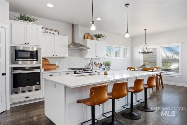 kitchen featuring white cabinets, an island with sink, decorative light fixtures, wall chimney range hood, and appliances with stainless steel finishes