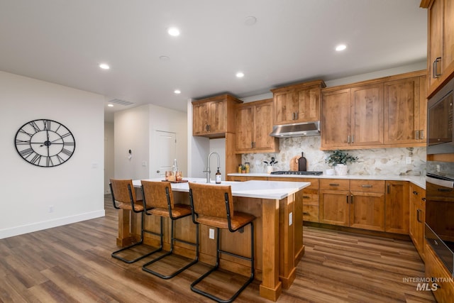 kitchen with dark wood-style floors, a kitchen island with sink, decorative backsplash, stainless steel gas stovetop, and under cabinet range hood