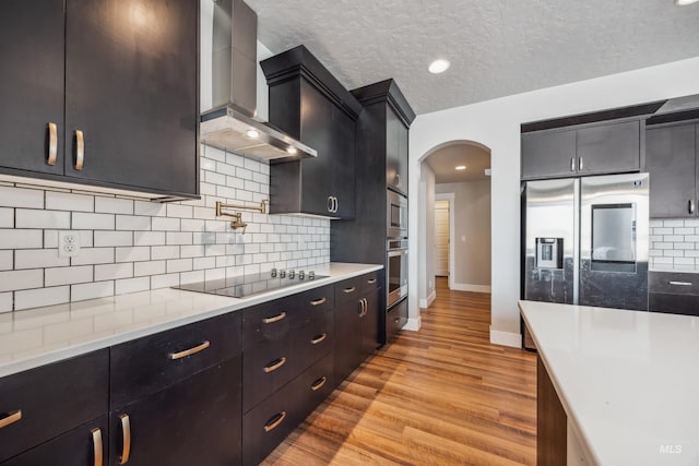 kitchen featuring light wood-style flooring, stainless steel appliances, arched walkways, wall chimney exhaust hood, and light countertops