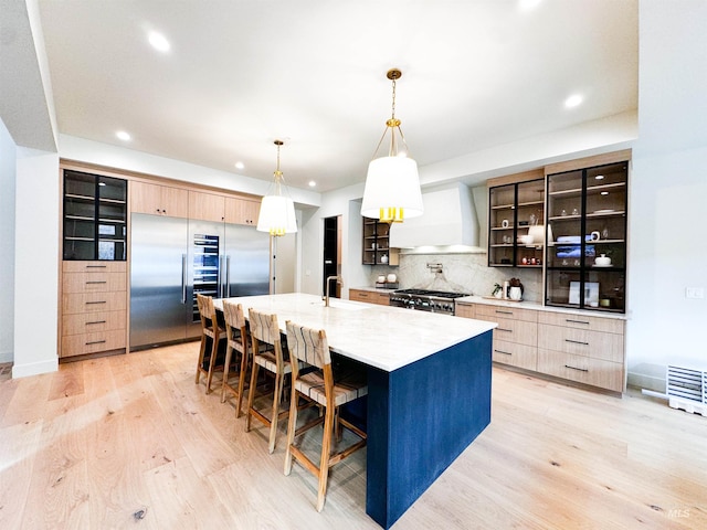 kitchen featuring light brown cabinets, custom range hood, hanging light fixtures, and a center island with sink