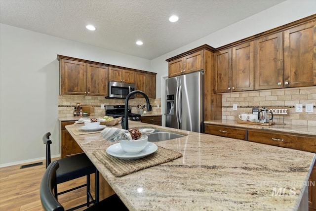 kitchen featuring appliances with stainless steel finishes, a sink, light stone countertops, light wood finished floors, and a kitchen island with sink