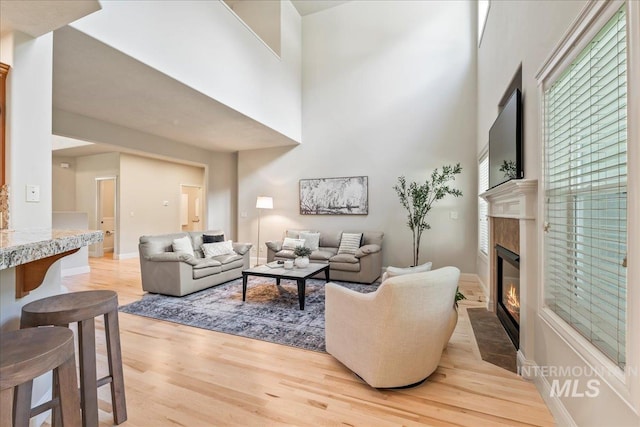 living room featuring light wood finished floors, a fireplace with flush hearth, a towering ceiling, and baseboards