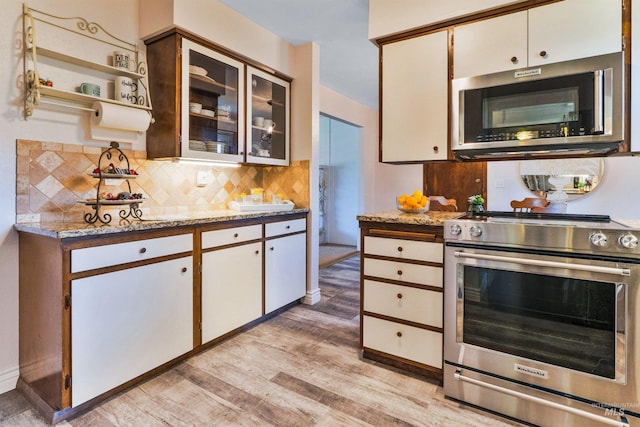 kitchen featuring stainless steel appliances, light wood-style flooring, tasteful backsplash, light stone countertops, and glass insert cabinets