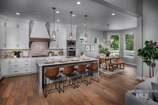 kitchen with a center island with sink, appliances with stainless steel finishes, light wood-type flooring, custom range hood, and pendant lighting