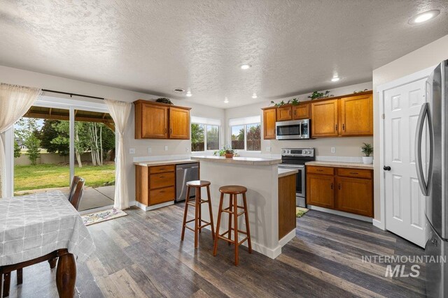 12339 W Foxhaven St, Star, ID 83669 - kitchen featuring appliances with stainless steel finishes, brown cabinetry, dark wood-style flooring, a kitchen island, and a textured ceiling