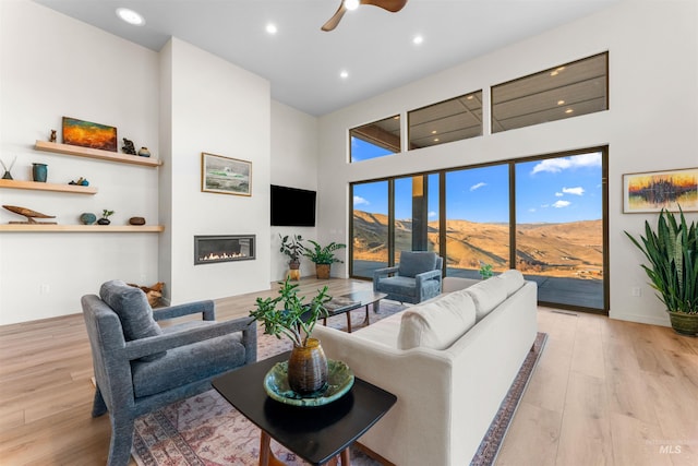 living room featuring light wood finished floors, ceiling fan, a high ceiling, and a glass covered fireplace