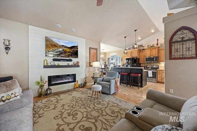 living room with light wood-type flooring, vaulted ceiling, arched walkways, a glass covered fireplace, and a textured ceiling