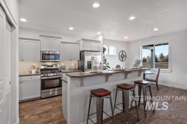 kitchen with a center island with sink, dark hardwood / wood-style floors, appliances with stainless steel finishes, a kitchen breakfast bar, and backsplash