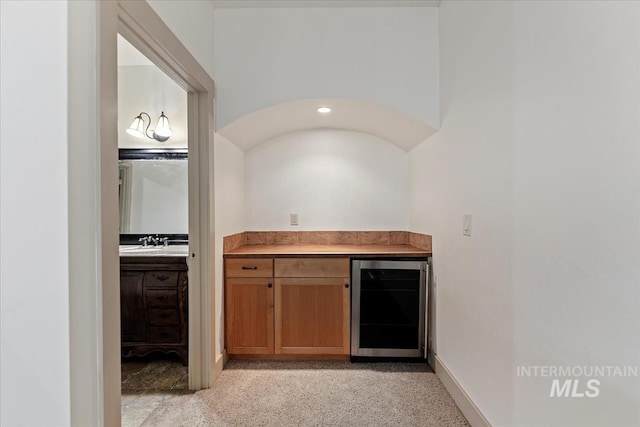 kitchen featuring brown cabinetry, baseboards, a sink, wine cooler, and light carpet