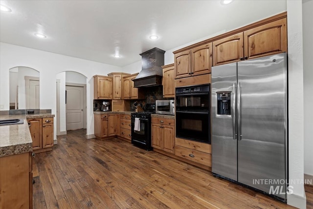 kitchen with premium range hood, brown cabinets, black appliances, backsplash, and dark wood finished floors