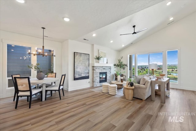 living room featuring light wood-type flooring, ceiling fan with notable chandelier, a textured ceiling, high vaulted ceiling, and a stone fireplace