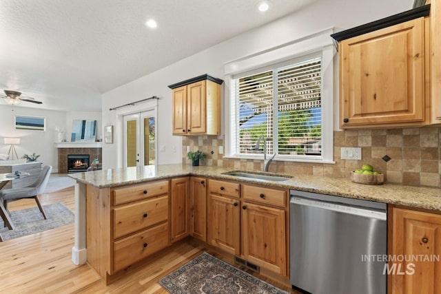 kitchen featuring stainless steel dishwasher, a sink, a tile fireplace, a peninsula, and light wood-style flooring