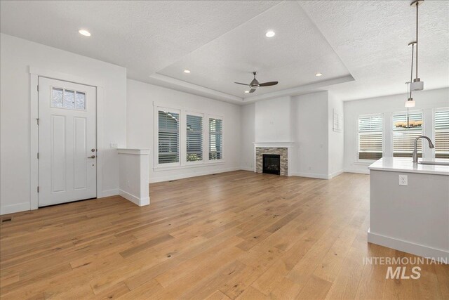 unfurnished living room featuring a stone fireplace, light hardwood / wood-style floors, a textured ceiling, a tray ceiling, and ceiling fan