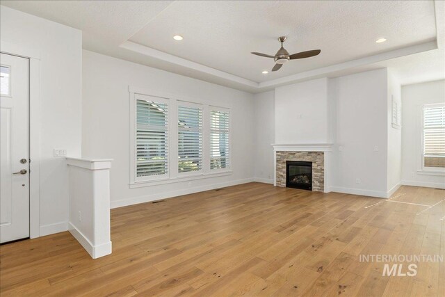 unfurnished living room with light wood-type flooring, a textured ceiling, a tray ceiling, ceiling fan, and a stone fireplace