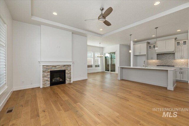 unfurnished living room with sink, a stone fireplace, light wood-type flooring, a tray ceiling, and ceiling fan