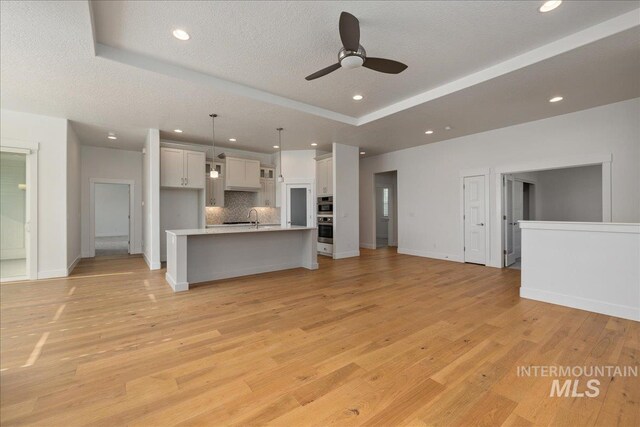 unfurnished living room with sink, light hardwood / wood-style flooring, a textured ceiling, a tray ceiling, and ceiling fan