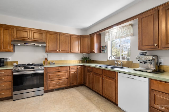 kitchen featuring under cabinet range hood, a sink, stainless steel gas range, white dishwasher, and brown cabinetry