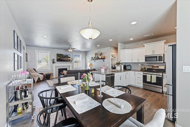 dining area featuring a ceiling fan, visible vents, wood finished floors, a warm lit fireplace, and recessed lighting