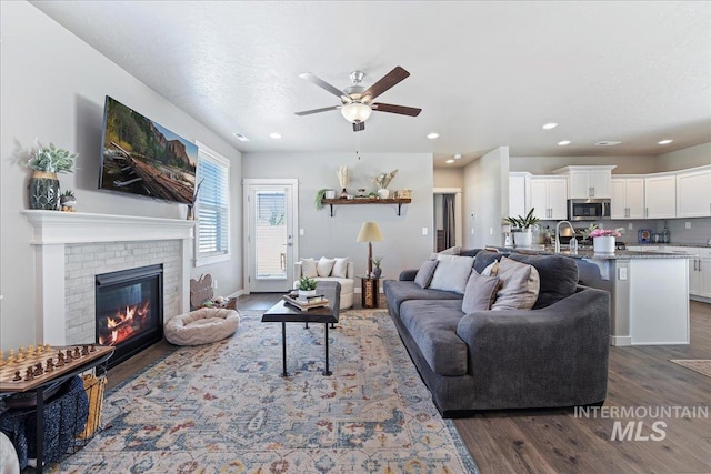 living room with a brick fireplace, a ceiling fan, recessed lighting, and dark wood-type flooring