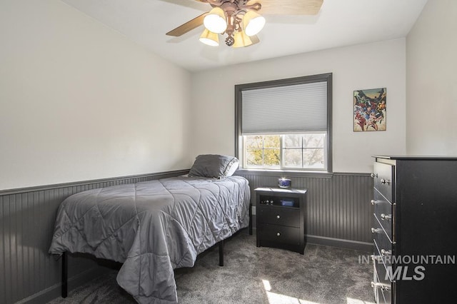 bedroom featuring a ceiling fan, a wainscoted wall, dark carpet, and wooden walls