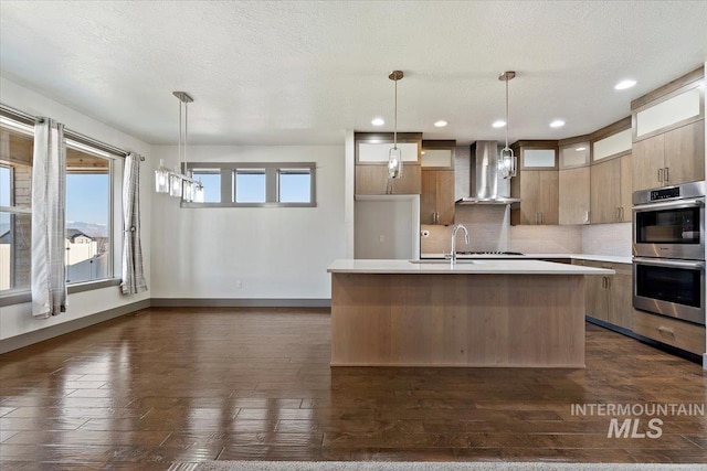 kitchen featuring pendant lighting, double oven, wall chimney range hood, and an island with sink