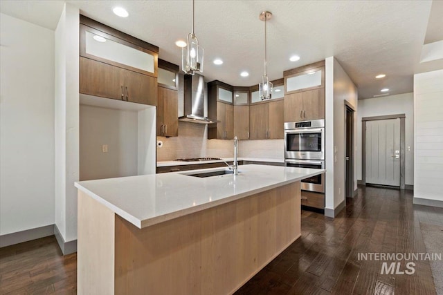 kitchen with wall chimney range hood, an island with sink, hanging light fixtures, and double oven