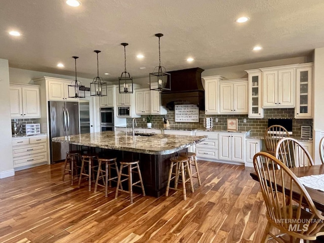 kitchen featuring wood finished floors, a large island, custom range hood, appliances with stainless steel finishes, and a kitchen bar