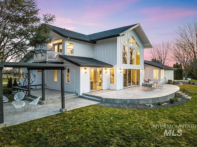 back of house at dusk with roof with shingles, a patio, a balcony, and a lawn