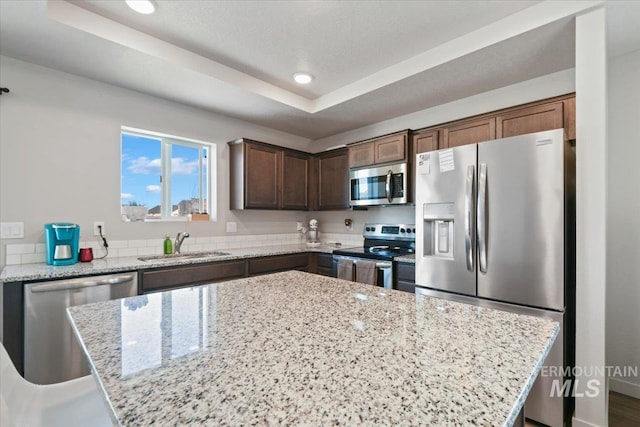 kitchen with light stone counters, stainless steel appliances, a sink, a center island, and a raised ceiling