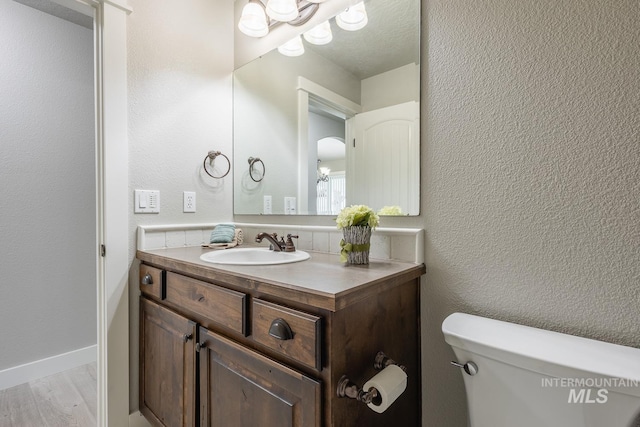 bathroom with vanity, wood finished floors, baseboards, toilet, and a textured wall