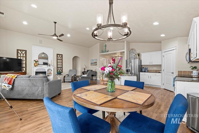 dining area featuring arched walkways, ceiling fan with notable chandelier, light wood finished floors, and a warm lit fireplace