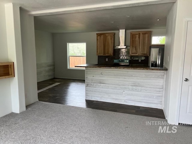 kitchen with stainless steel refrigerator with ice dispenser, wall chimney range hood, dark countertops, dark colored carpet, and brown cabinetry